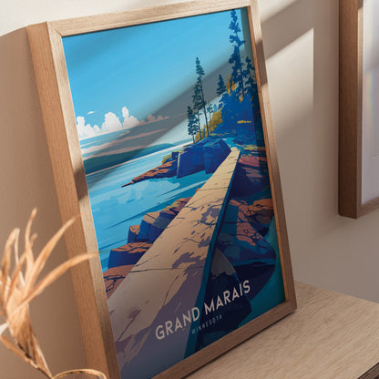 Framed artwork depicting a vibrant landscape of Grand Marais, Minnesota, featuring a rocky shoreline and pine trees under a clear blue sky. The poster rests against a light-colored wall.