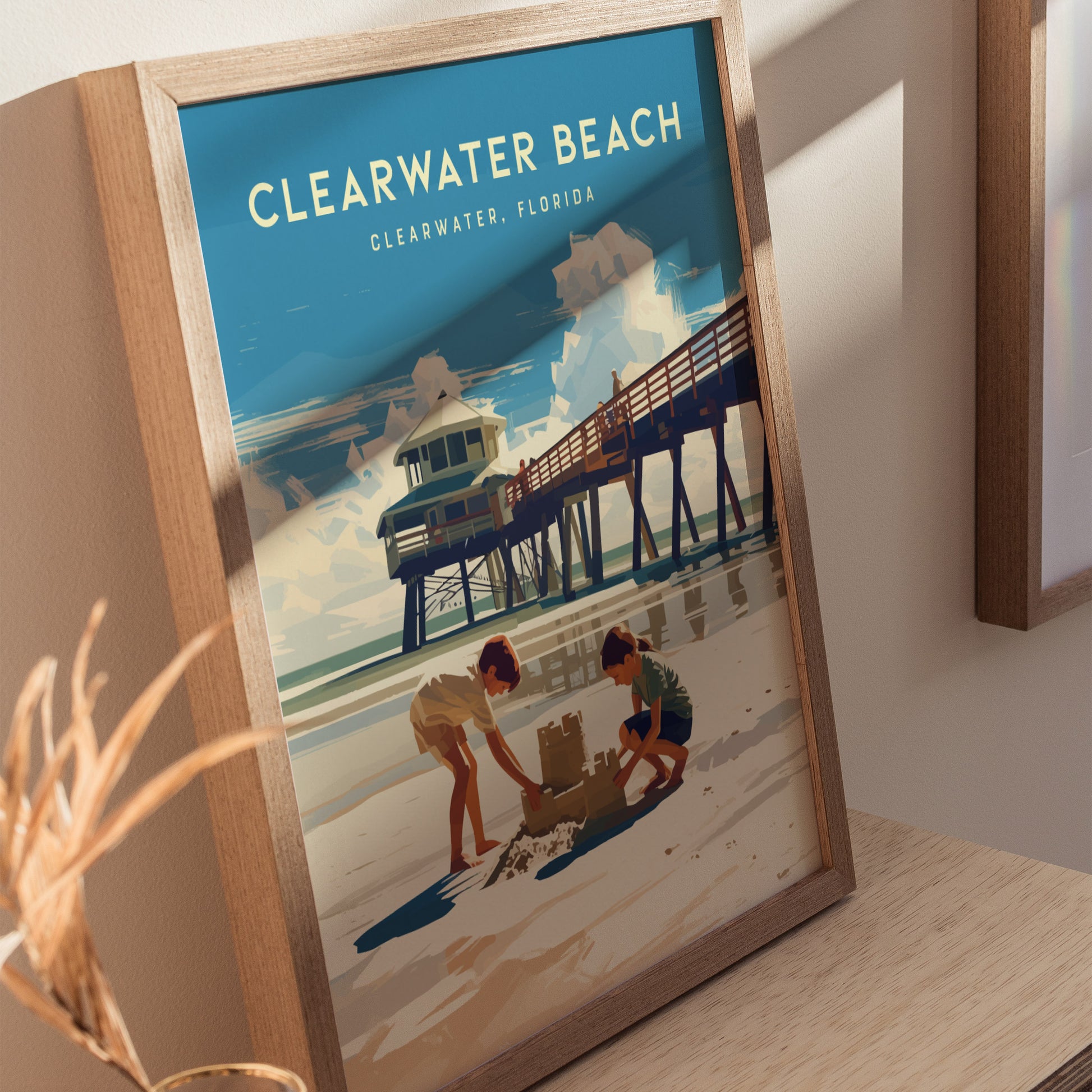 Framed artwork of Clearwater Beach shows two children building a sandcastle on the beach near a wooden pier, under a bright blue sky.