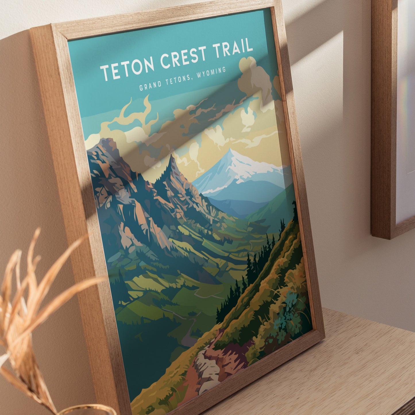 Framed poster of the Teton Crest Trail featuring colorful mountains and valleys under a cloudy sky, displayed on a light wooden surface.