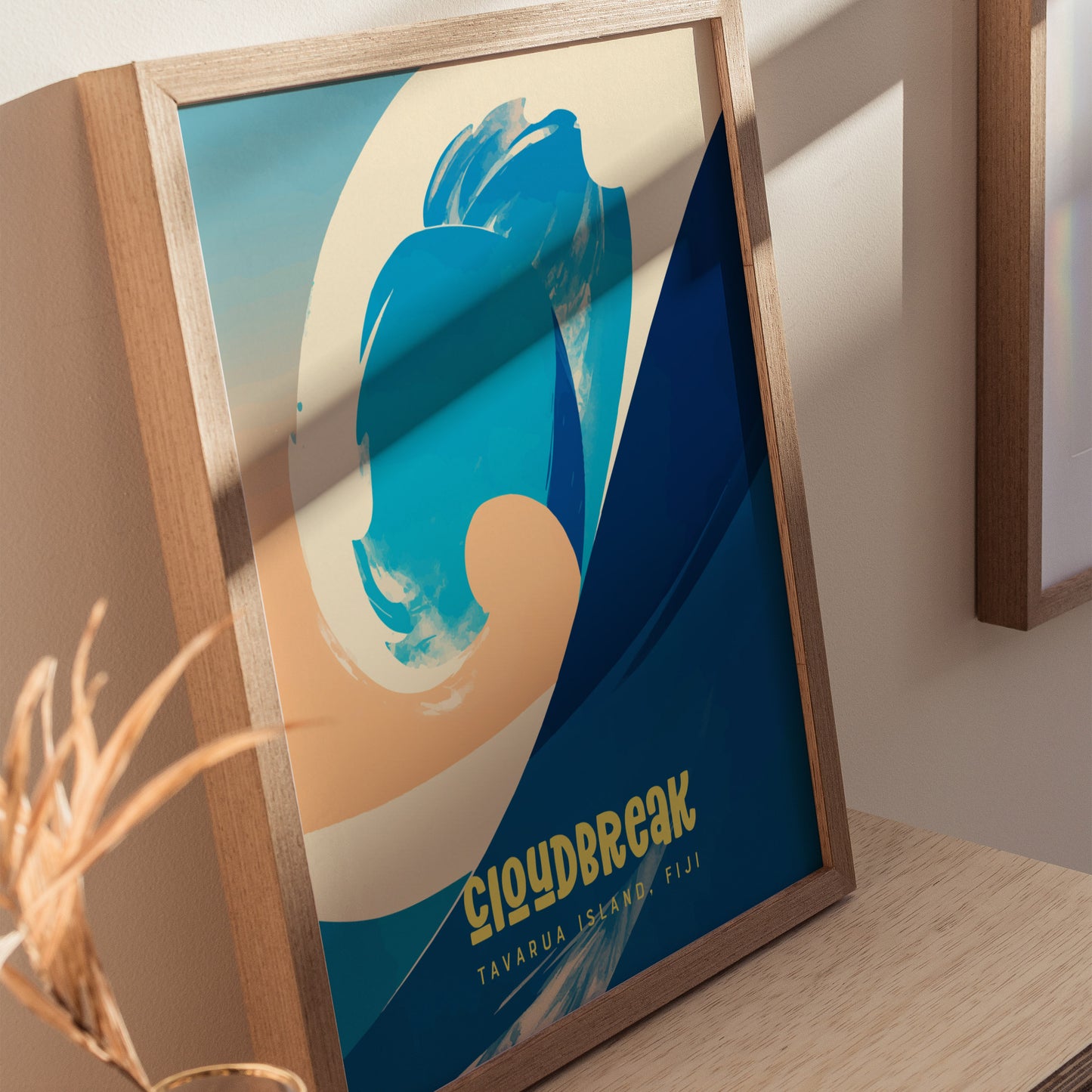 A framed poster featuring abstract wave artwork in shades of blue and beige, with the words Cloudbreak Tavarua Island, Fiji at the bottom, displayed on a wooden surface.