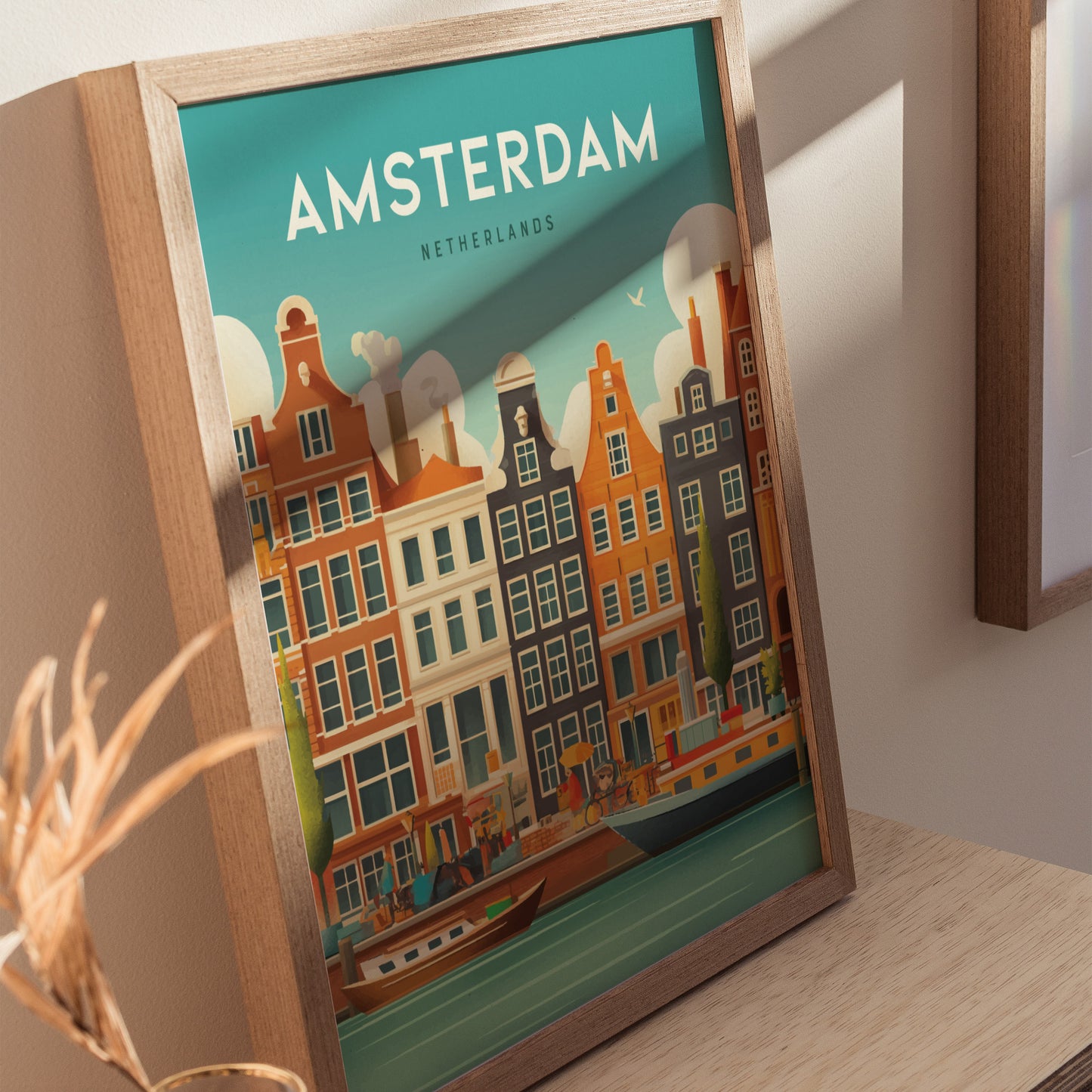 Framed artwork depicting colorful Amsterdam canal houses under a blue sky, with the text AMSTERDAM NETHERLANDS at the top. The print rests on a wooden surface in soft lighting.
