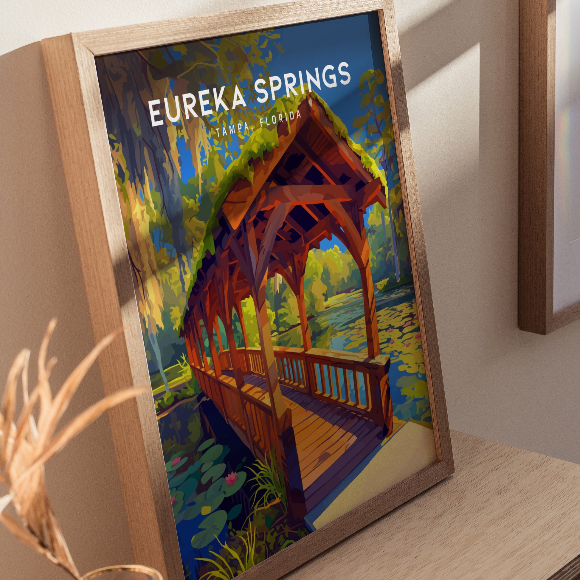 Framed artwork of Eureka Springs in Tampa, Florida, showcasing a wooden bridge over a lily-filled stream, surrounded by lush greenery and Spanish moss.