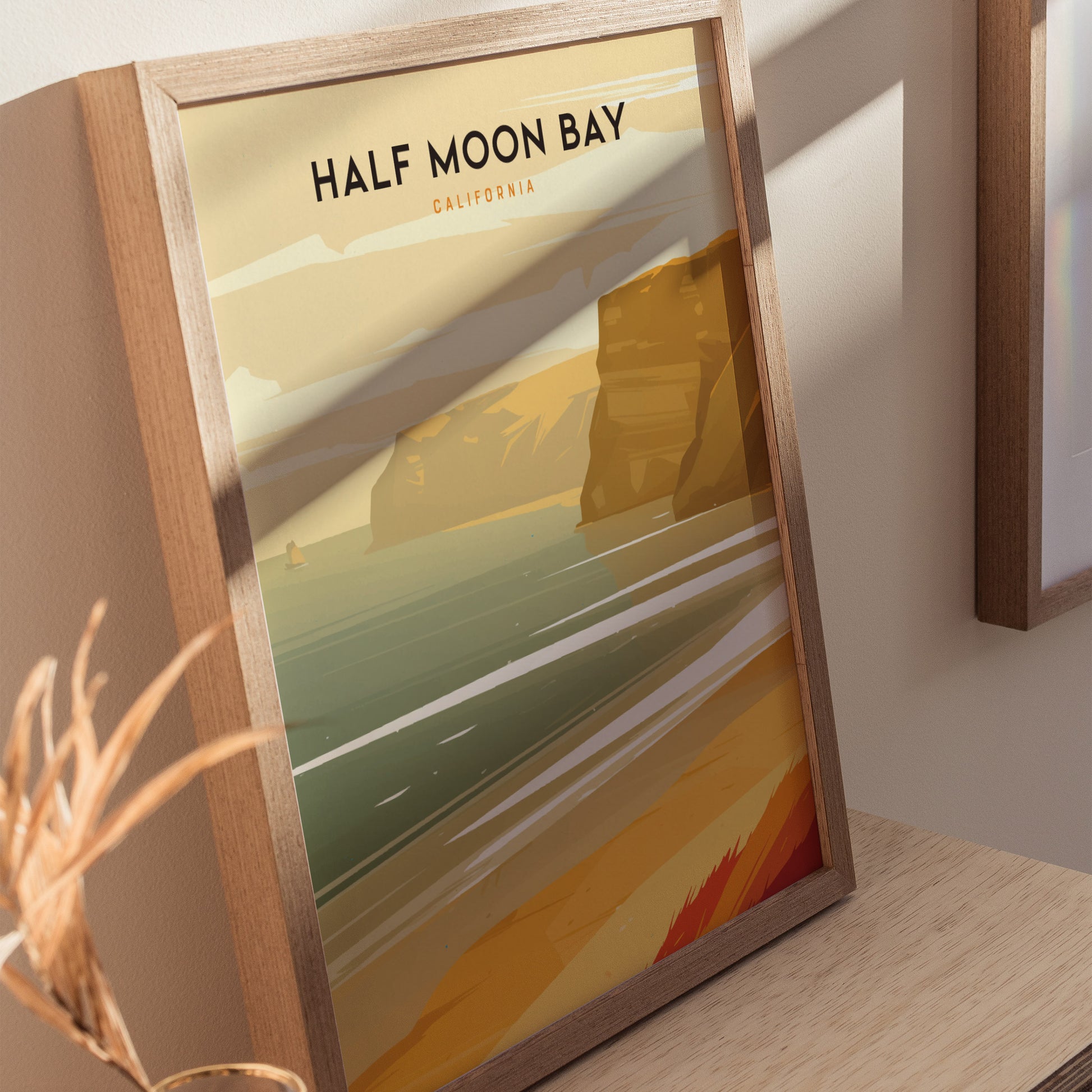 Framed poster depicting Half Moon Bay with stylized golden cliffs and ocean waves, set against a beige wall. Sunlight casts shadows on the artwork and wooden surface.