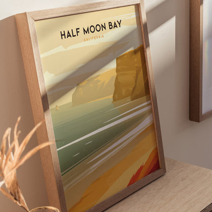 Framed poster depicting Half Moon Bay with stylized golden cliffs and ocean waves, set against a beige wall. Sunlight casts shadows on the artwork and wooden surface.