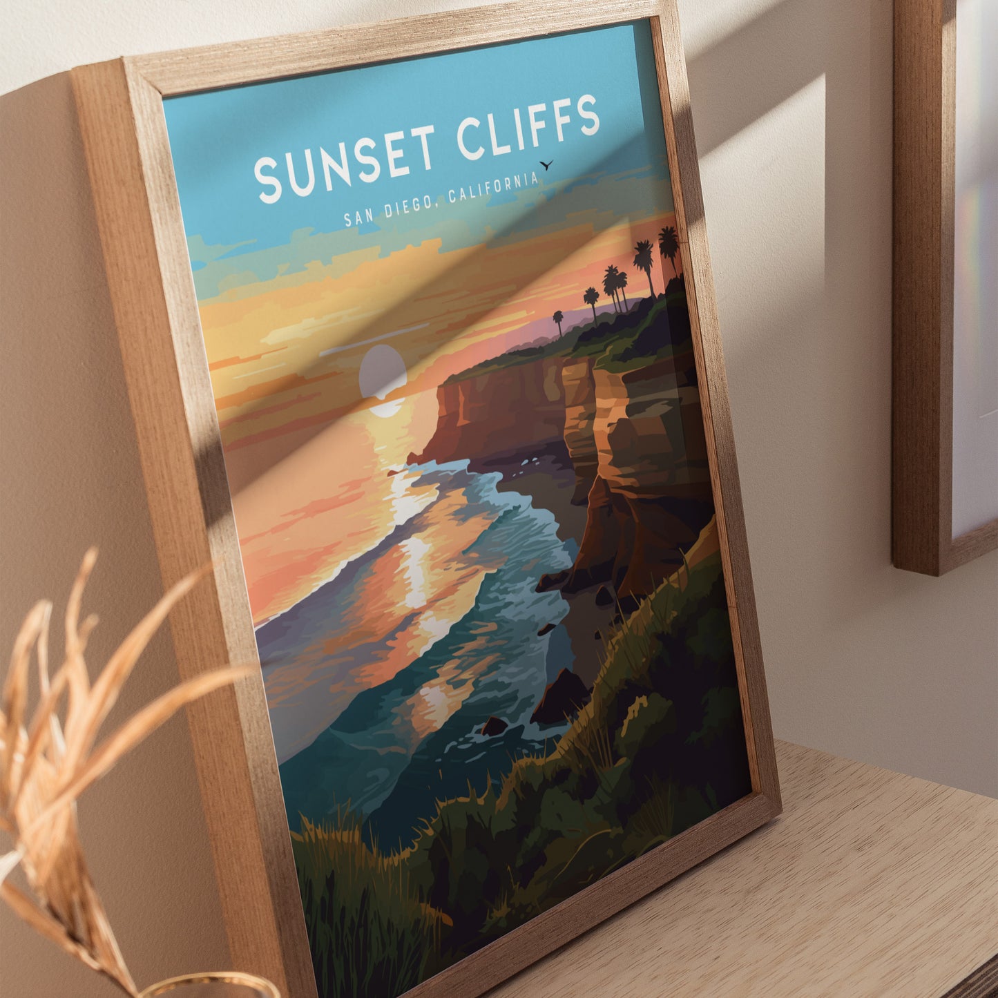 Framed artwork titled Sunset Cliffs depicts a vibrant sunset over ocean waves and cliffs in San Diego, California. The scene includes silhouetted palm trees and a glowing horizon.