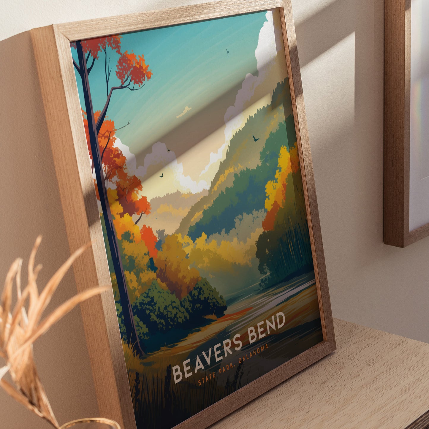 Framed poster of Beavers Bend State Park features vibrant autumn foliage, rolling hills, and a serene river under a blue sky. Displayed on a wooden surface with soft lighting.