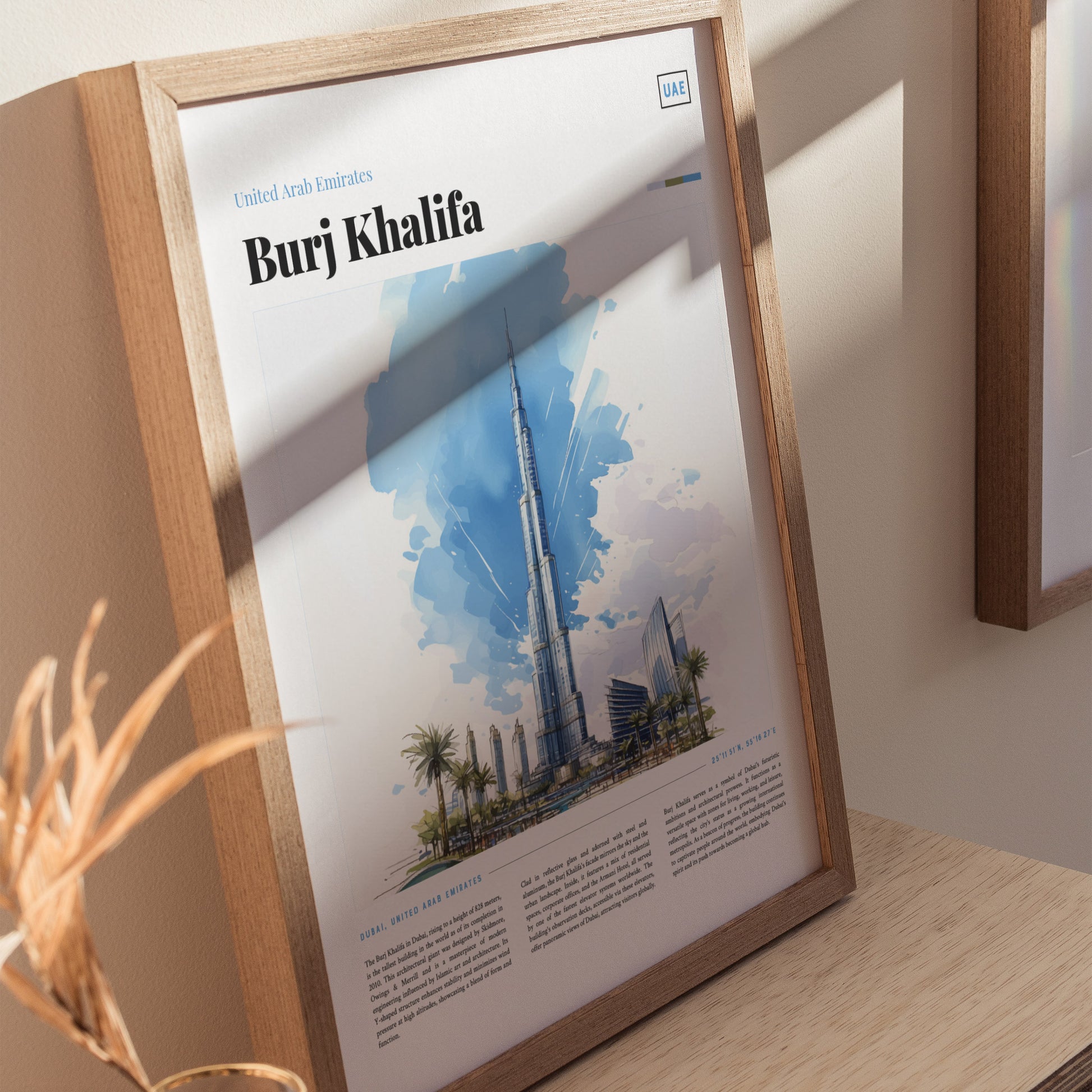 A framed poster featuring a watercolor illustration of the Burj Khalifa against a blue sky, displayed on a wooden surface with soft shadows and decorative elements.