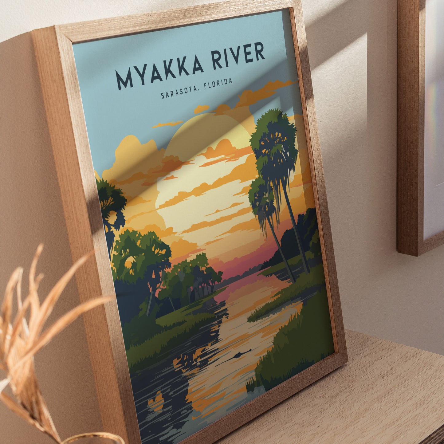 Framed Myakka River State Park poster featuring a colorful sunset over a river with palm trees and clouds, resting on a wooden surface with soft lighting.