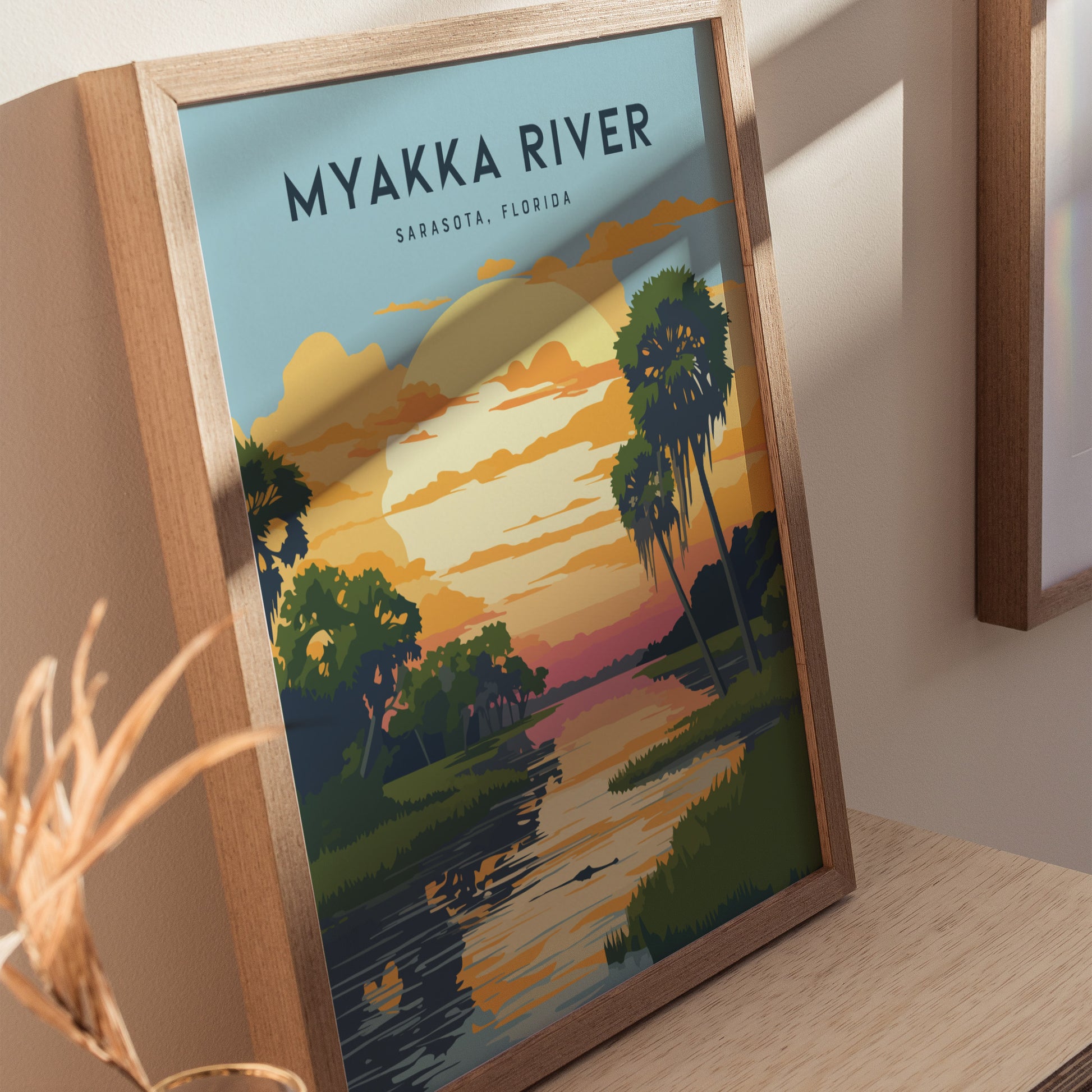 Framed Myakka River State Park poster featuring a colorful sunset over a river with palm trees and clouds, resting on a wooden surface with soft lighting.