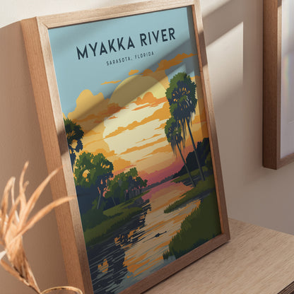 Framed Myakka River State Park poster featuring a colorful sunset over a river with palm trees and clouds, resting on a wooden surface with soft lighting.