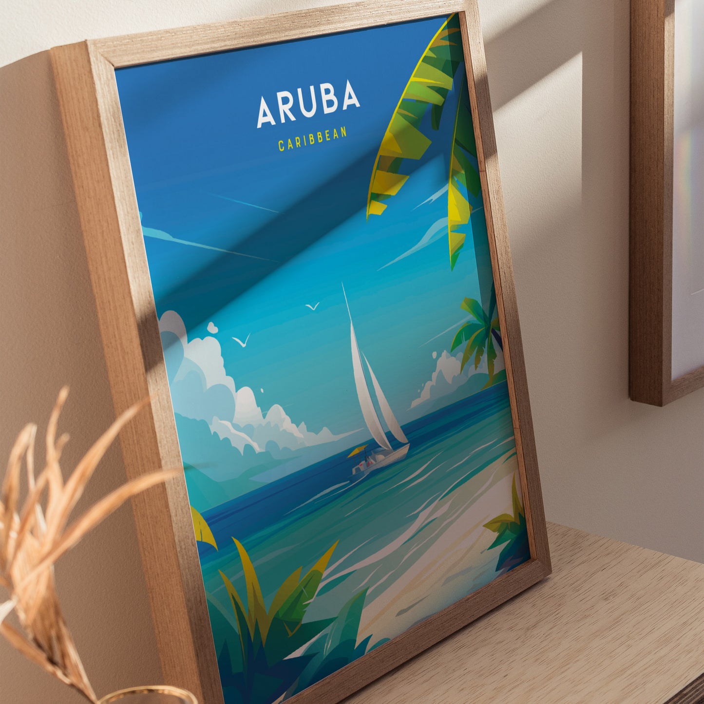 A framed poster depicts a vibrant Caribbean scene with a sailboat on turquoise waters, lush palm leaves, and Aruba Caribbean text at the top. Sunlight casts shadows on the wall.