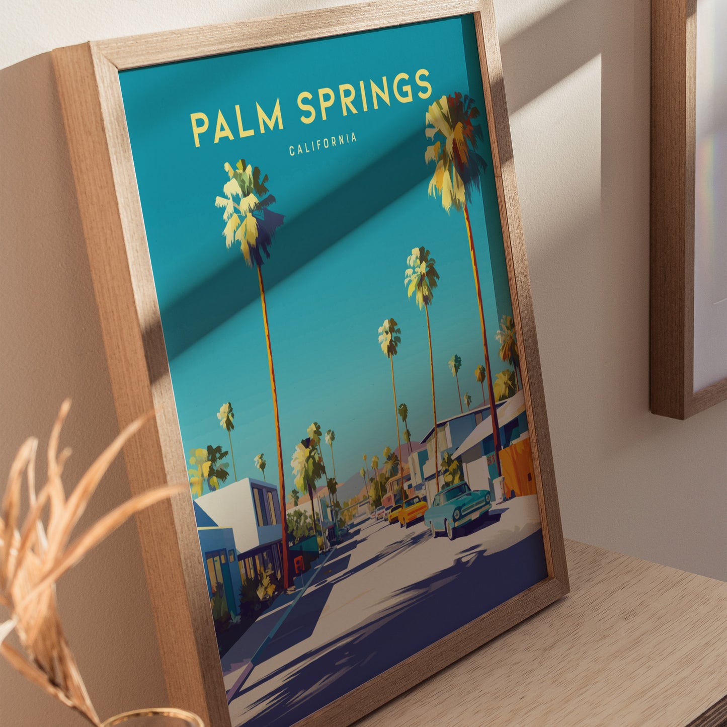 Framed Palm Springs poster featuring palm trees, classic cars, and mid-century houses under a clear blue sky. The vibrant artwork is displayed on a wooden surface with soft lighting.