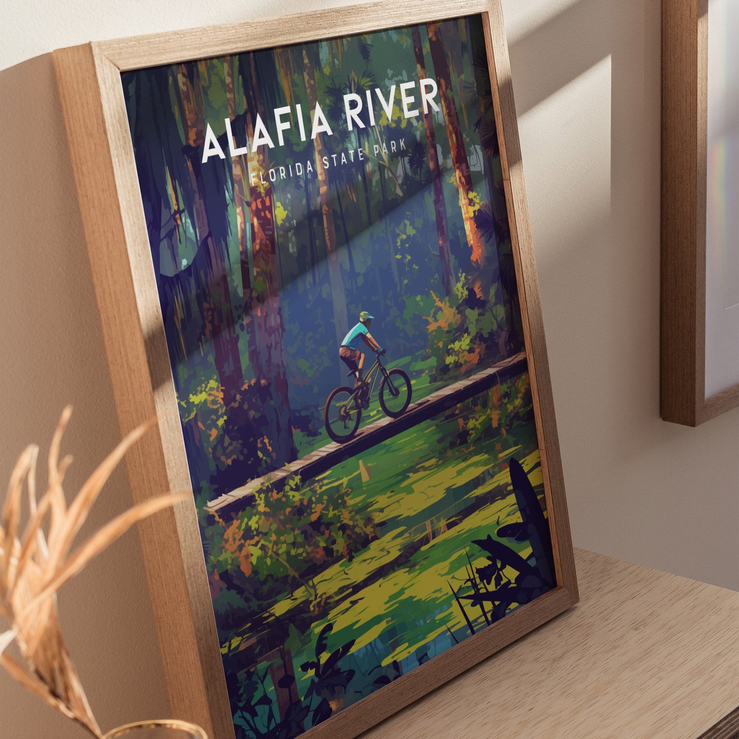 Framed poster of Alafia River State Park depicting a cyclist crossing a wooden bridge surrounded by lush forest and a calm creek, set against a light-colored wall.
