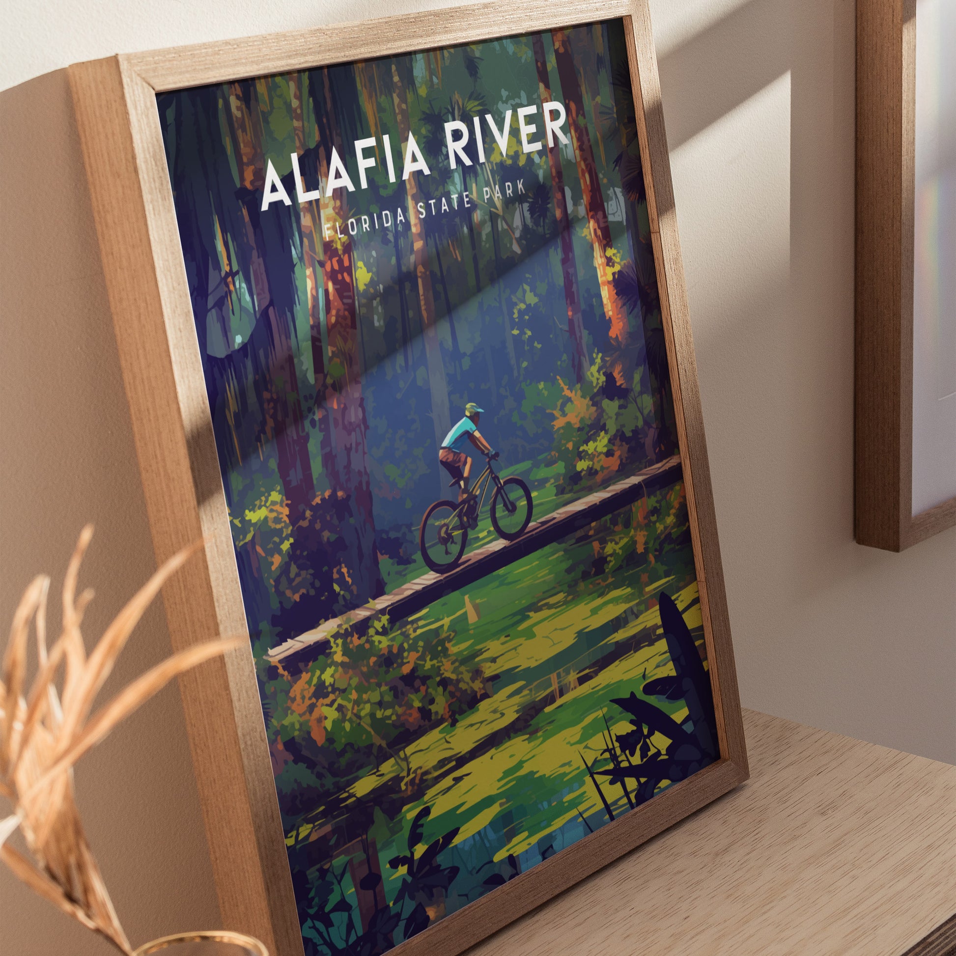 Framed poster of Alafia River State Park depicting a cyclist crossing a wooden bridge surrounded by lush forest and a calm creek, set against a light-colored wall.