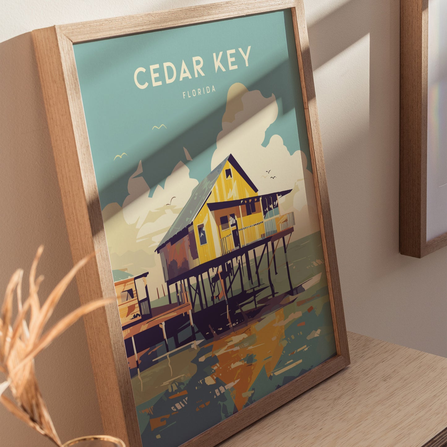 Framed Cedar Key poster depicting a colorful stilt house over coastal waters, with a backdrop of clouds and flying birds, placed on a wooden surface.