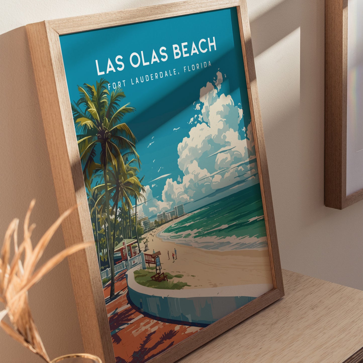Framed Las Olas Beach poster featuring palm trees, sandy beach, and ocean waves under a blue sky with fluffy clouds, displayed on a wooden surface with soft shadows.