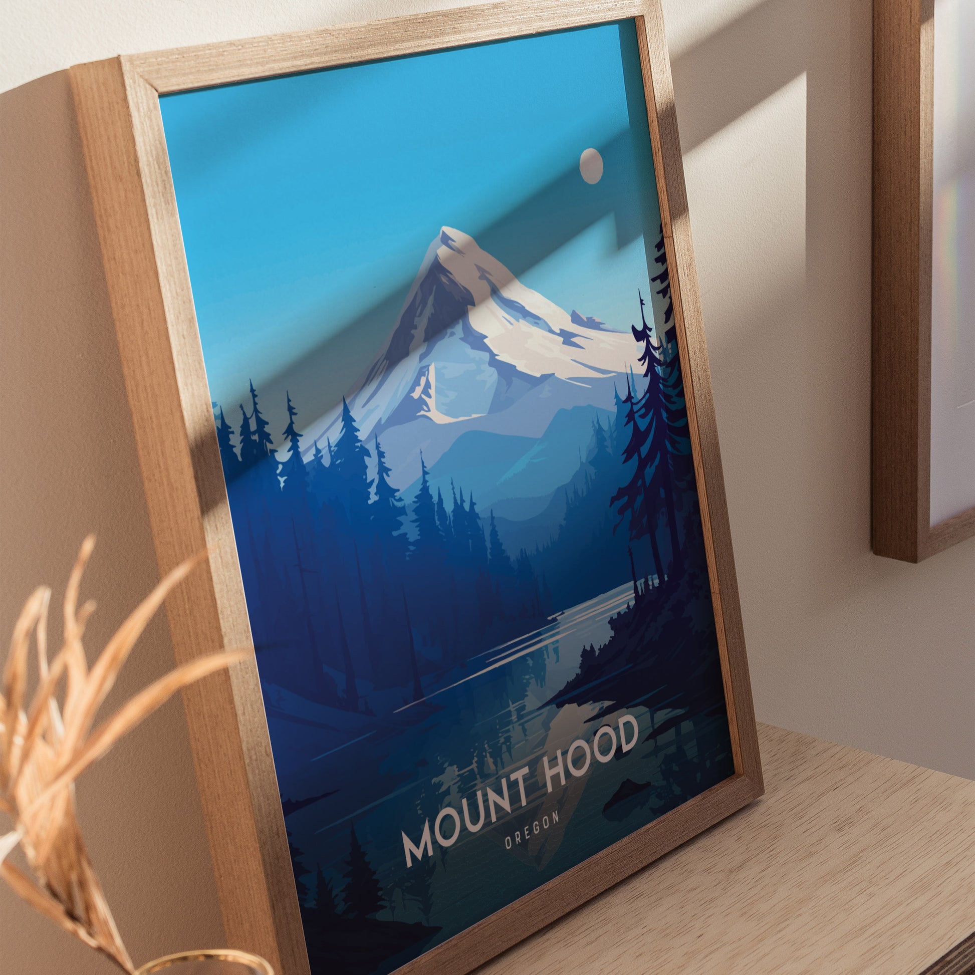 A framed poster of Mount Hood features a snow-capped peak, forested foothills, and a serene blue sky. It rests on a wooden surface with soft shadows.