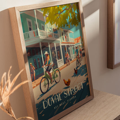 A framed poster depicts a vibrant scene on Duval Street, Key West, with cyclists, pedestrians, and roosters under leafy trees. The artwork captures a lively, tropical atmosphere.