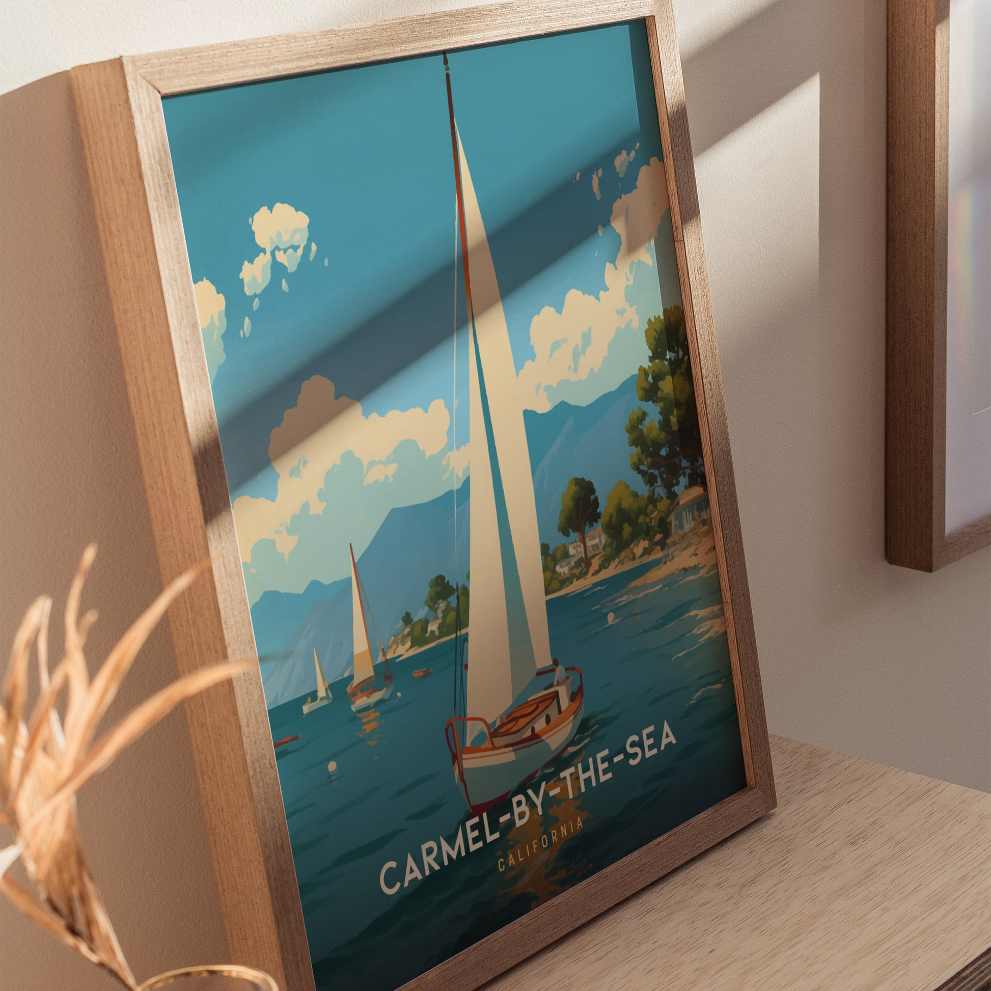 Framed poster featuring a sailboat on a serene blue sea with fluffy clouds and distant mountains, labeled Carmel-by-the-Sea California, displayed on a light wooden surface.