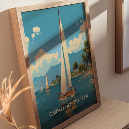 Framed poster featuring a sailboat on a serene blue sea with fluffy clouds and distant mountains, labeled Carmel-by-the-Sea California, displayed on a light wooden surface.