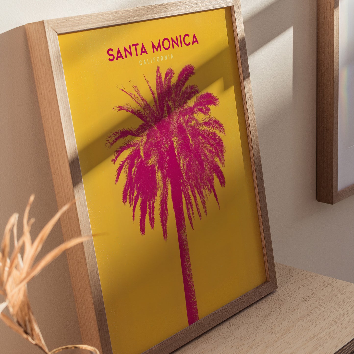 Framed artwork featuring a vibrant pink palm tree against a yellow background with Santa Monica California text. The poster is displayed on a light wooden surface.