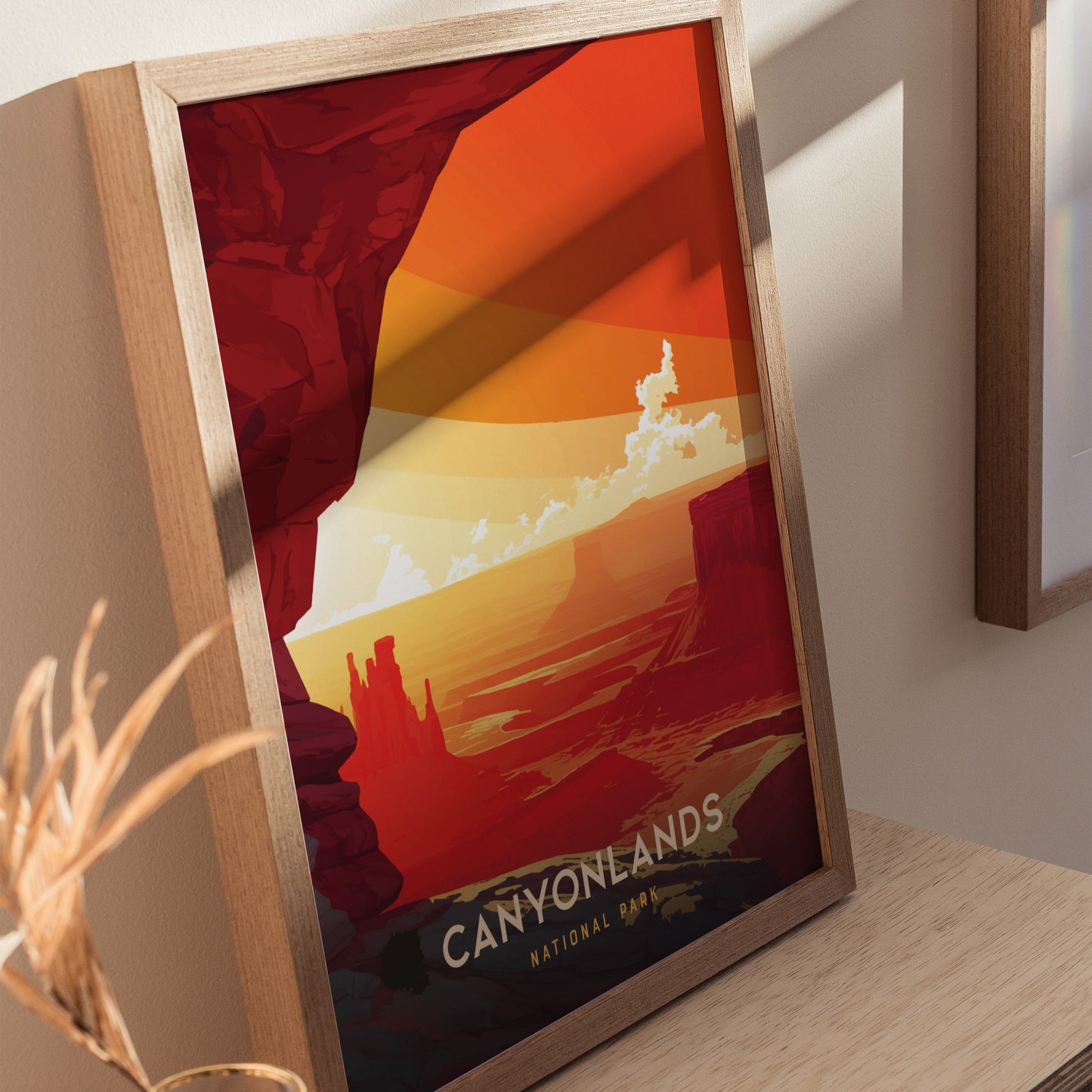 A framed art print depicting Canyonlands National Park with vibrant orange and red hues, showcasing dramatic desert canyons and mesas. The print rests on a wooden surface.