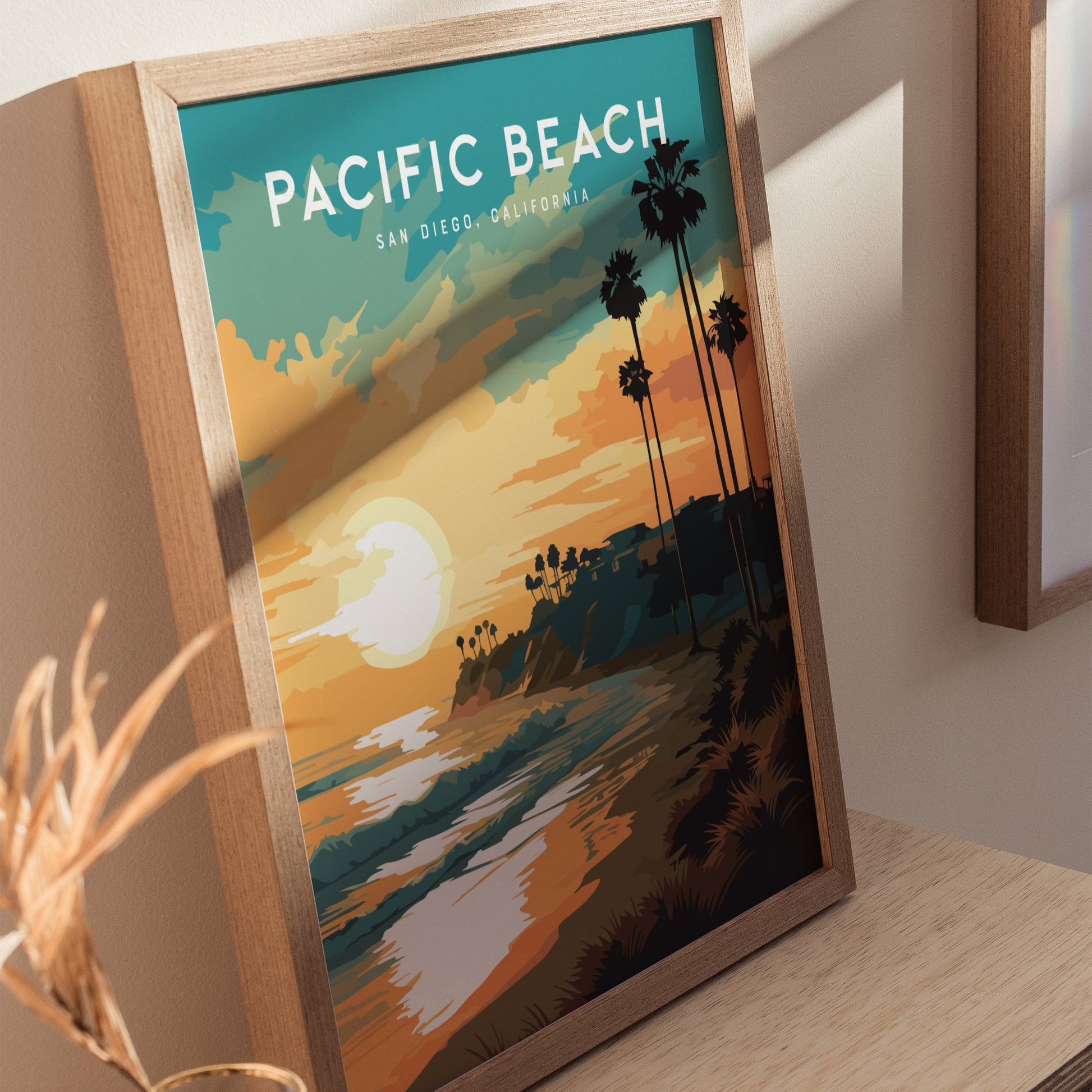 The image displays a framed Pacific Beach San Diego poster with a vibrant sunset scene, showcasing palm trees silhouetted against an orange and teal sky over the ocean. The poster rests on a wooden surface, adding warmth to the setting.