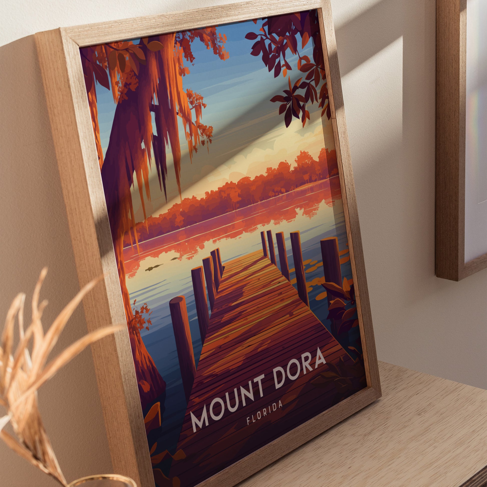 A framed poster of Mount Dora, Florida, depicts a scenic lakeside view with a wooden dock, vibrant orange trees, and a tranquil sunset. The artwork rests on a wooden surface.