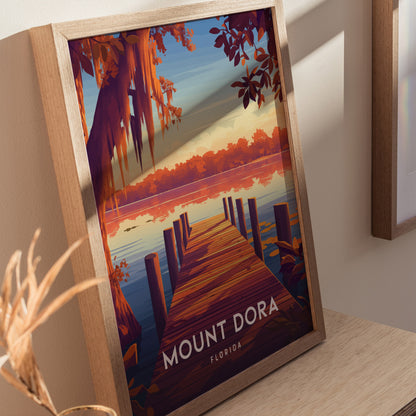 A framed poster of Mount Dora, Florida, depicts a scenic lakeside view with a wooden dock, vibrant orange trees, and a tranquil sunset. The artwork rests on a wooden surface.