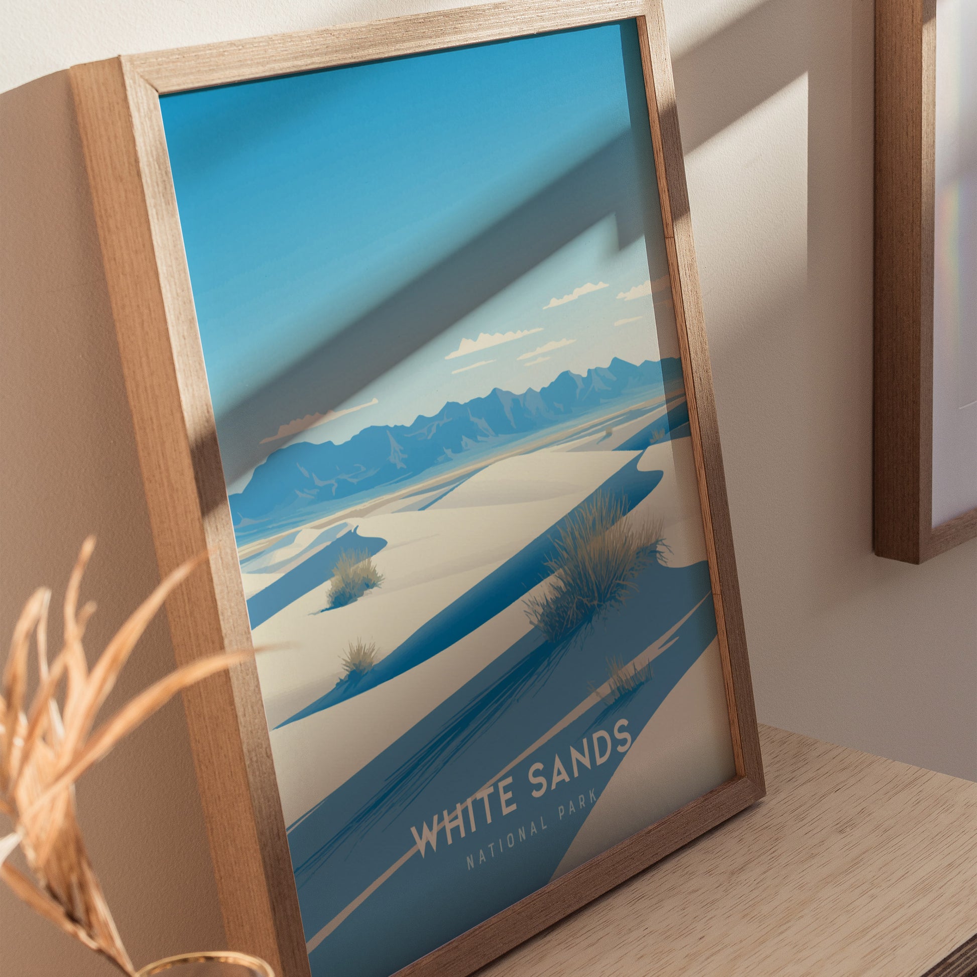 Framed artwork of White Sands National Park features blue skies, white dunes, and distant mountains, displayed on a wooden surface with natural light.