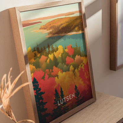 A framed poster featuring colorful autumn foliage in Lutsen, Minnesota, with vibrant hues of red, orange, and green trees against a blue lake backdrop, displayed on a wooden surface.