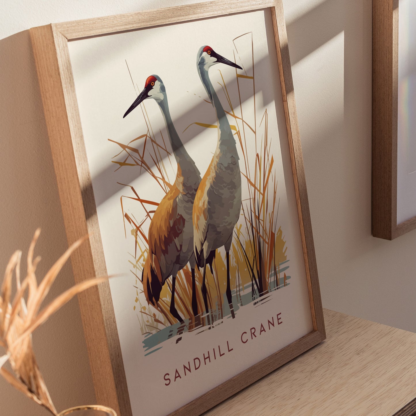 Framed artwork depicting two sandhill cranes standing amidst tall grasses, rendered in earthy tones. The image is displayed on a wooden surface with soft natural lighting.