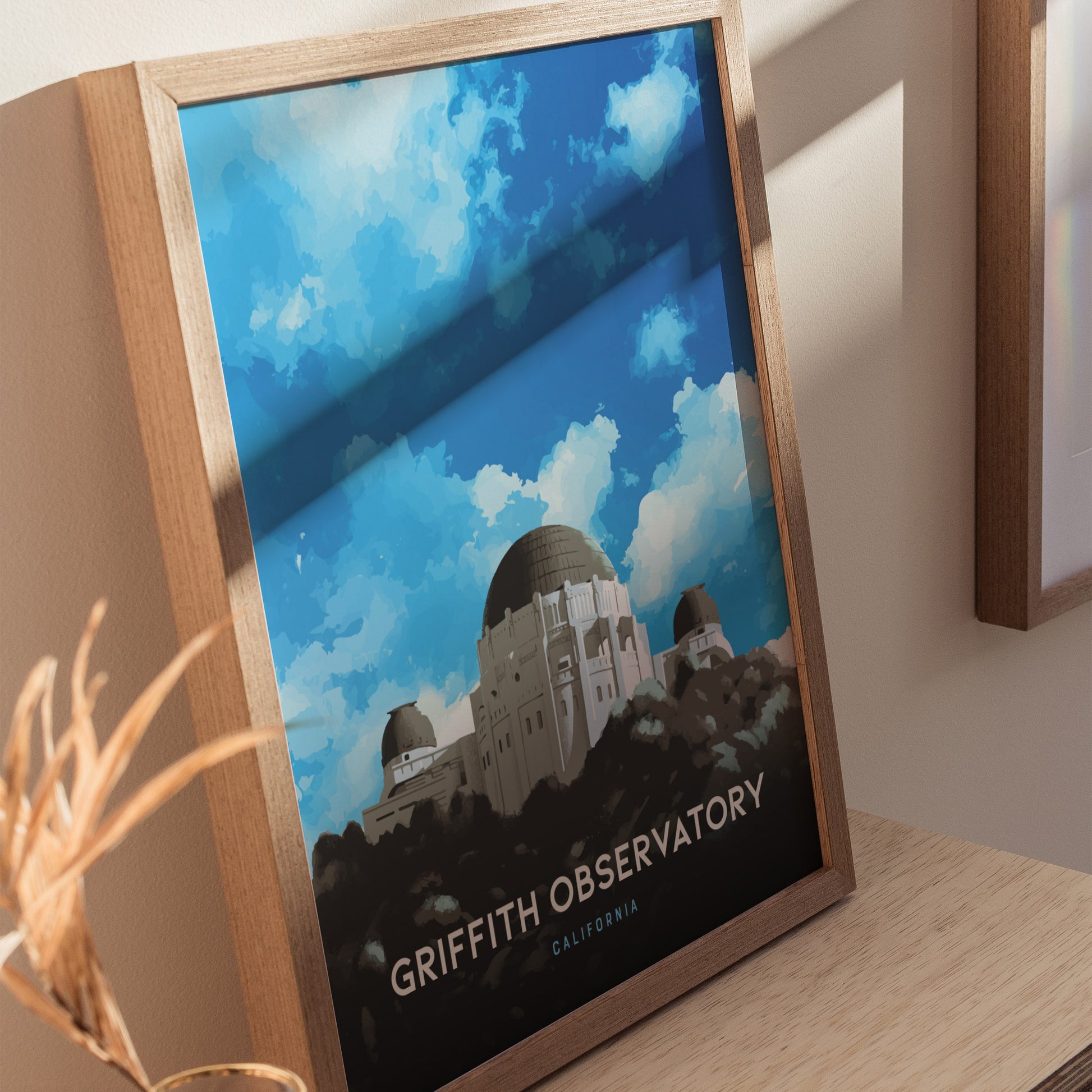 Framed Griffith Observatory poster featuring the observatorys domes against a vivid blue sky with clouds, displayed on a wooden surface with soft lighting.