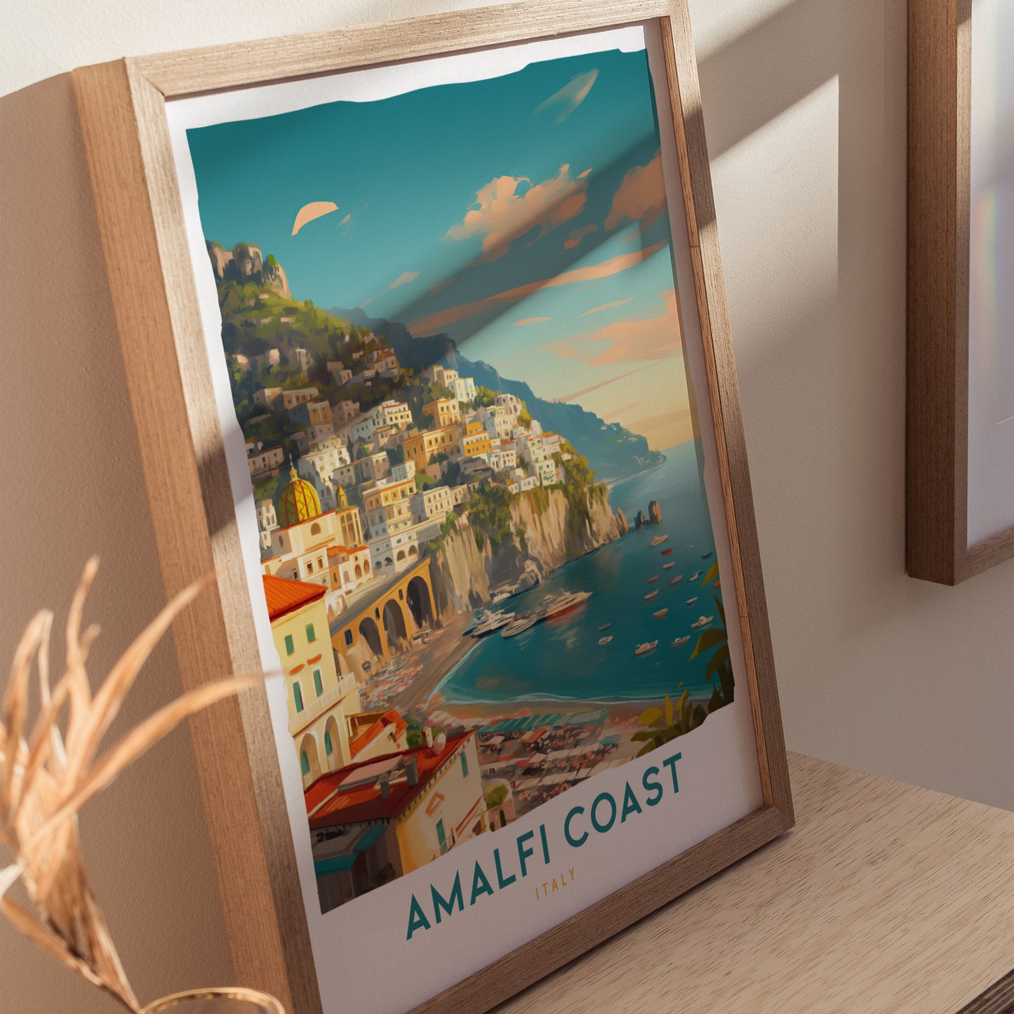 Framed artwork depicting the Amalfi Coast, featuring colorful cliffside buildings and a serene turquoise sea under a vibrant sky. The frame rests on a light wooden surface.