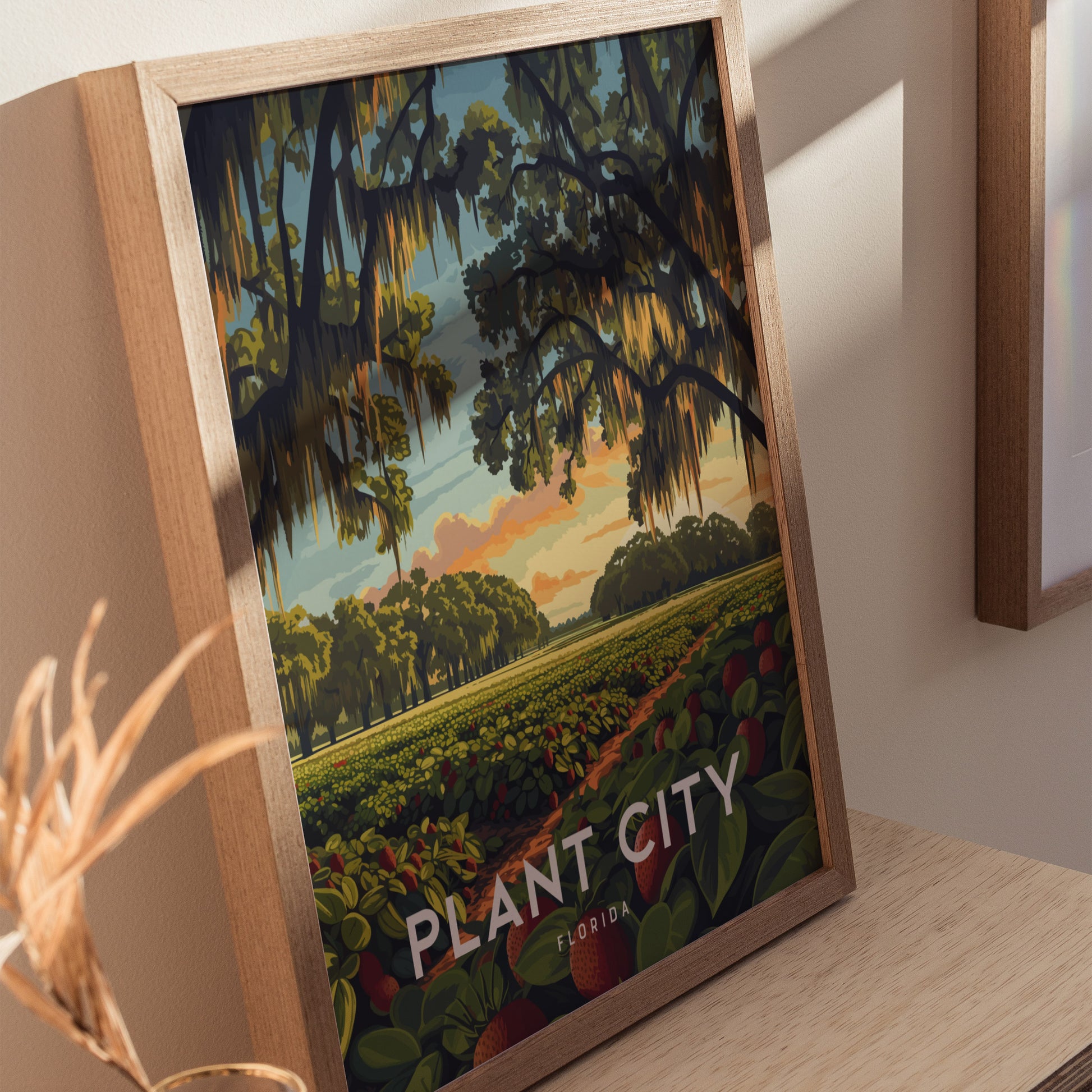 Framed artwork of Plant City, Florida, depicting a vibrant sunrise over strawberry fields with oak trees draped in Spanish moss. The poster rests on a wooden surface.