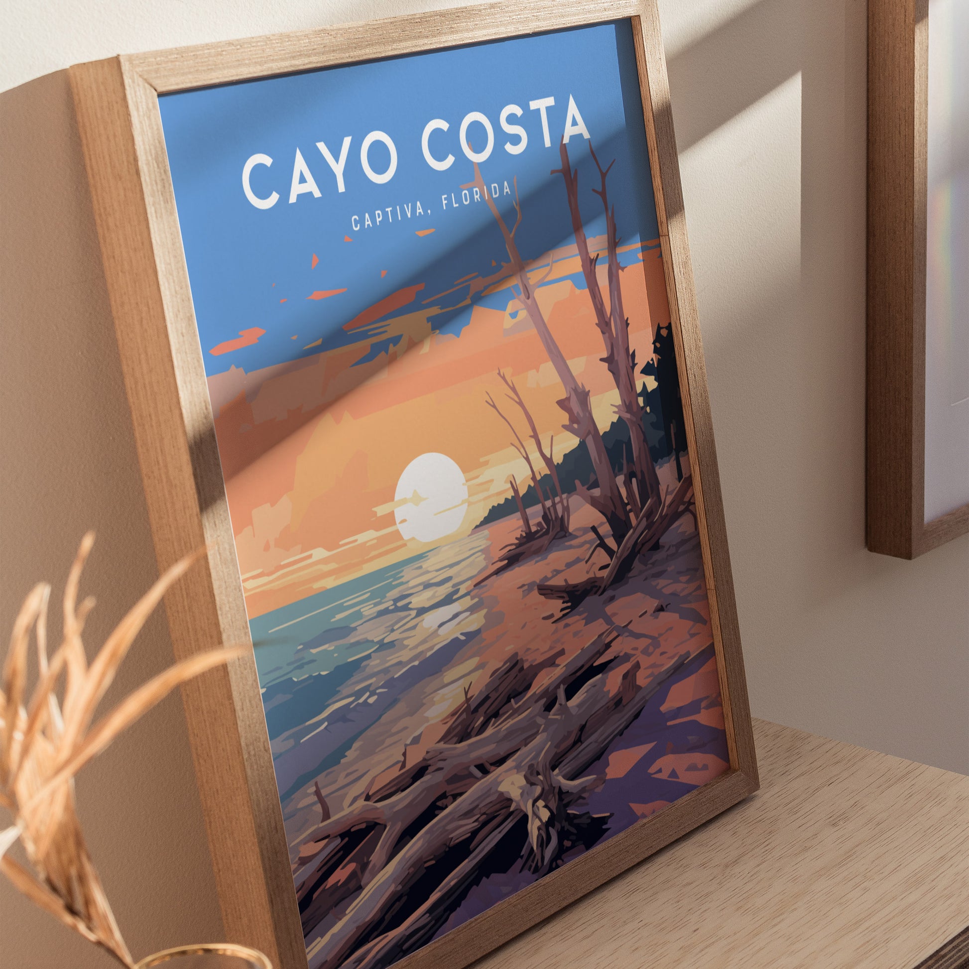A framed poster of Cayo Costa, Captiva, Florida, featuring a sunset over a beach with driftwood and vibrant colors, rests on a wooden surface.