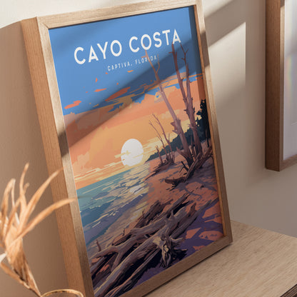A framed poster of Cayo Costa, Captiva, Florida, featuring a sunset over a beach with driftwood and vibrant colors, rests on a wooden surface.