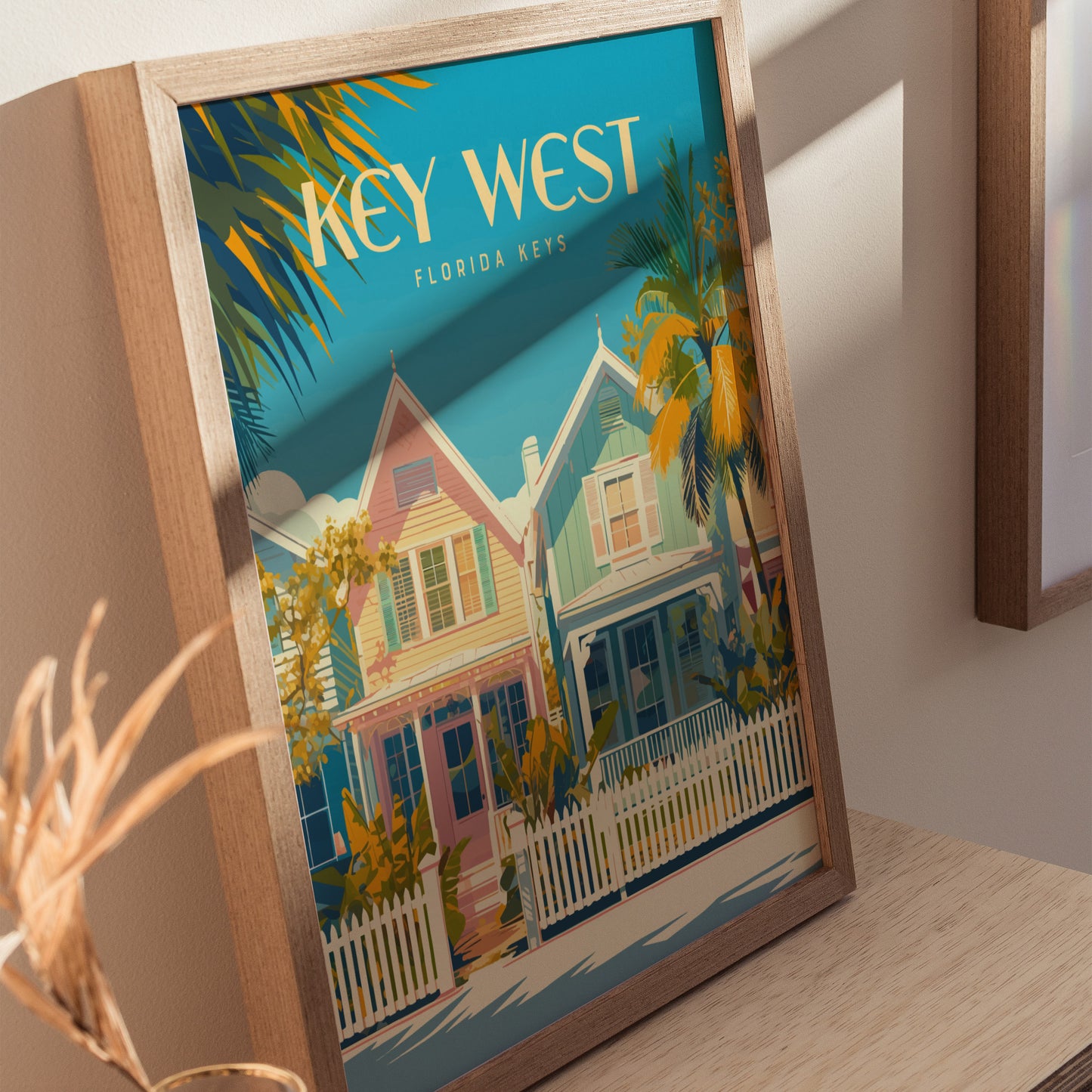Framed Key West poster featuring colorful Victorian-style houses, palm trees, and a bright blue sky. The artwork rests on a wooden surface, casting soft shadows.
