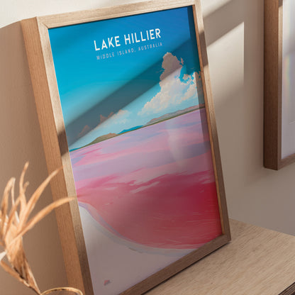 Framed poster of Lake Hillier features vibrant pink water contrasting with blue sky and clouds, displayed on a wooden surface. Perfect for adding color to any space.