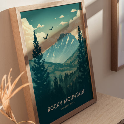 Framed poster featuring a scenic illustration of Rocky Mountain National Park, showcasing majestic peaks, pine trees, and birds in flight under a cloudy sky, resting on a wooden surface.