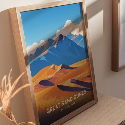 A framed poster of Great Sand Dunes National Park features vibrant golden dunes against blue mountains and a clear sky, resting on a wooden surface.