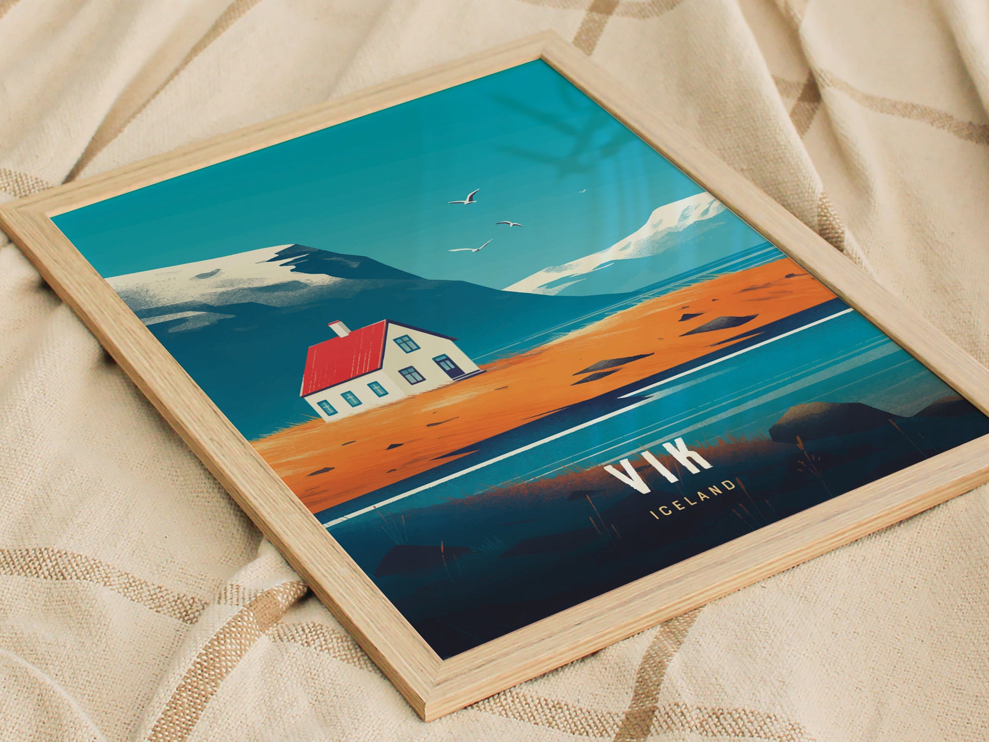 A framed poster featuring a stylized landscape with a house, red roof, and mountainous backdrop, titled Vik Iceland, resting on a soft, beige fabric surface.