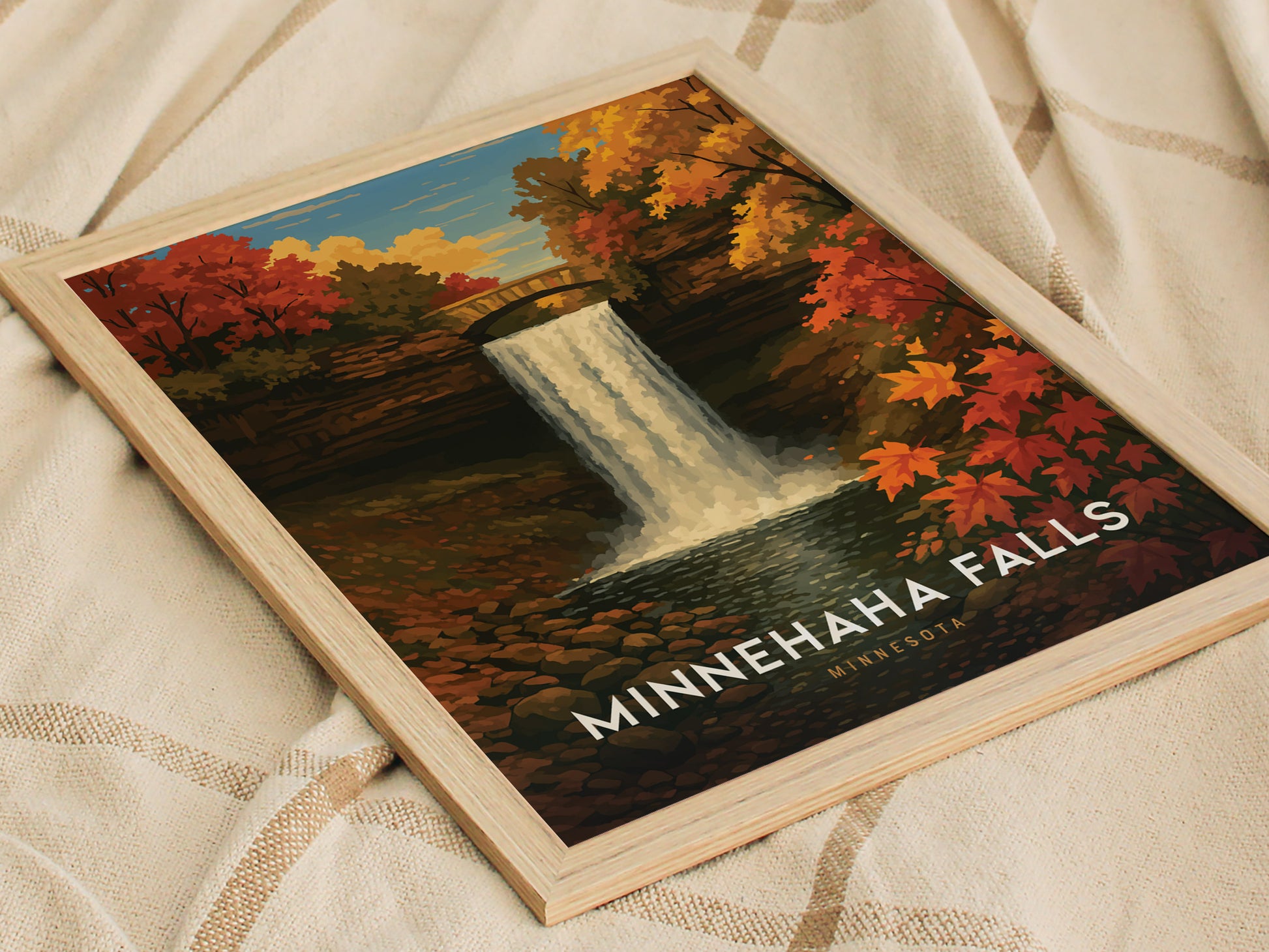 Framed poster of Minnehaha Falls, featuring a waterfall with vibrant autumn foliage and a stone bridge, displayed on a textured beige fabric background.