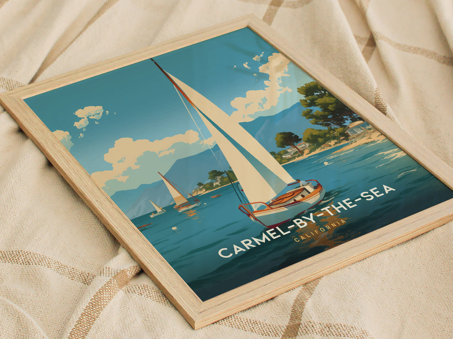 A framed poster depicting a sailboat on a serene blue ocean with a coastline and trees in the background, titled Carmel-by-the-Sea, California, rests on a beige fabric.