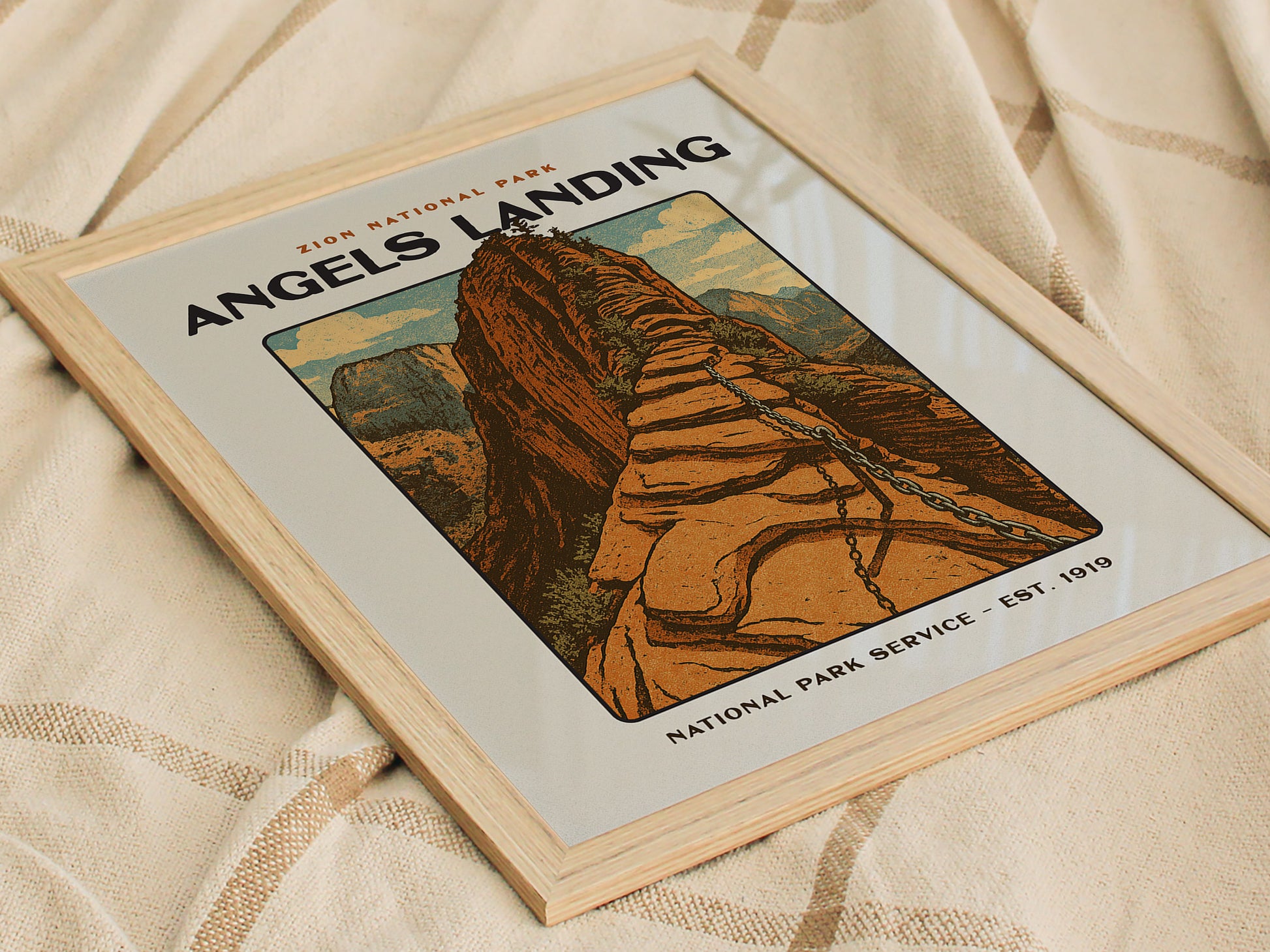 Framed vintage-style poster of Angels Landing in Zion National Park, featuring a bold block print design with steep sandstone formations and canyon walls, resting on a textured fabric surface.