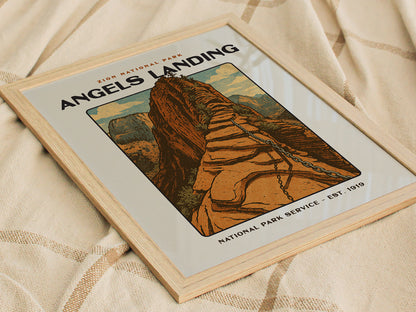 Framed vintage-style poster of Angels Landing in Zion National Park, featuring a bold block print design with steep sandstone formations and canyon walls, resting on a textured fabric surface.