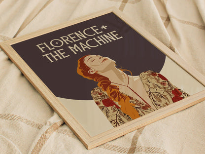 A framed Florence + The Machine poster featuring an illustrated woman with red hair in a floral robe, set against a dark background, lies on a beige fabric.