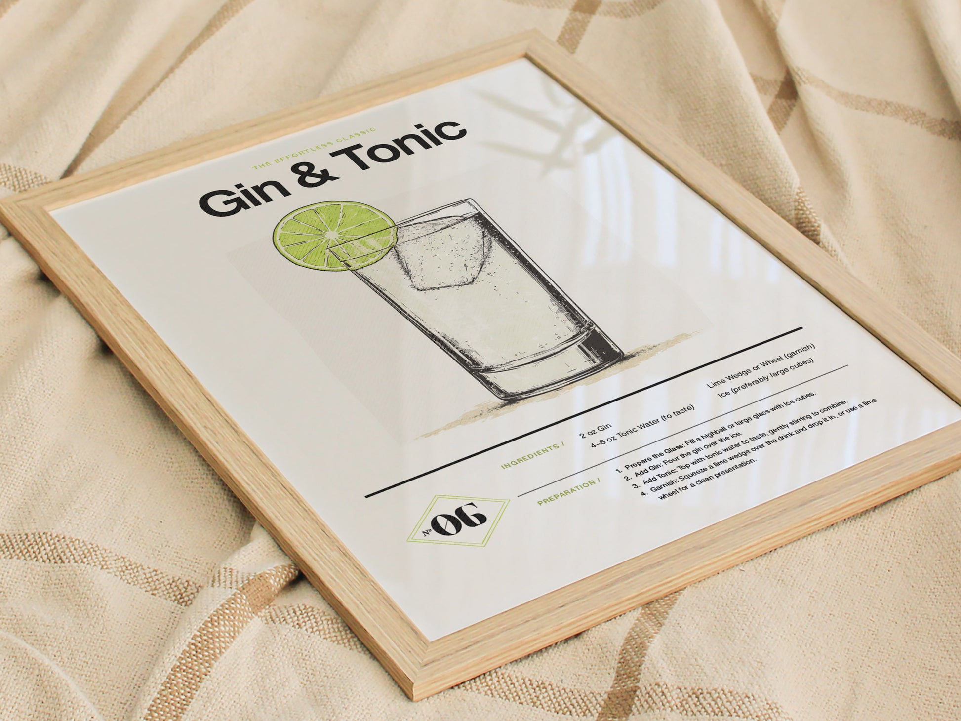 Framed poster with Gin & Tonic title, featuring a minimalist illustration of the drink with a lime slice garnish. Displayed on a soft, textured fabric background.