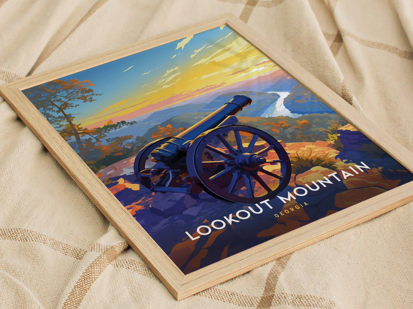A framed poster of Lookout Mountain, Georgia, featuring a historic cannon overlooking a scenic landscape with a colorful sunset sky, is placed on a soft, beige fabric.