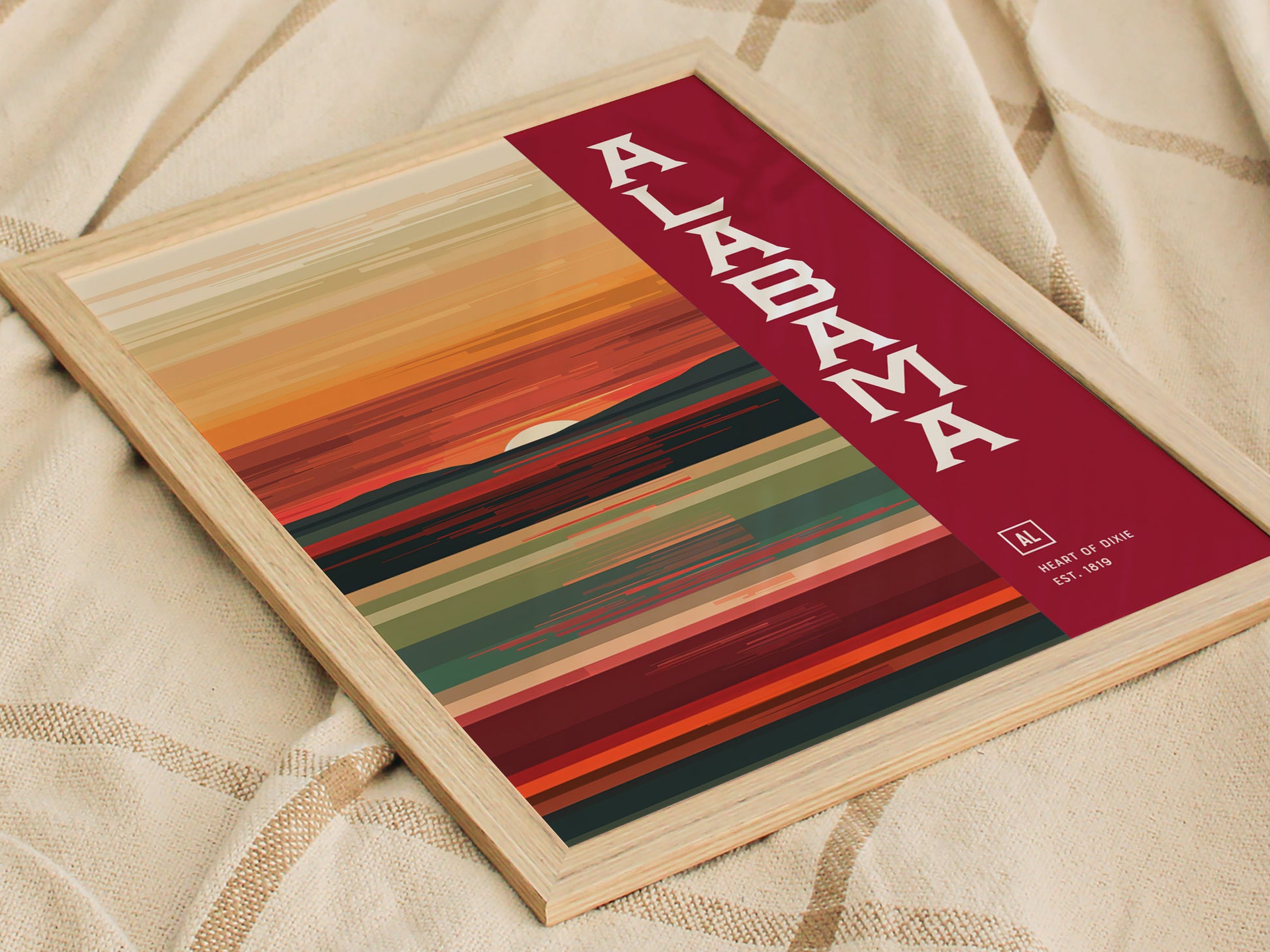 A framed abstract poster featuring warm auburn and crimson stripes with the word Alabama boldly displayed on a red background, resting on a textured fabric.